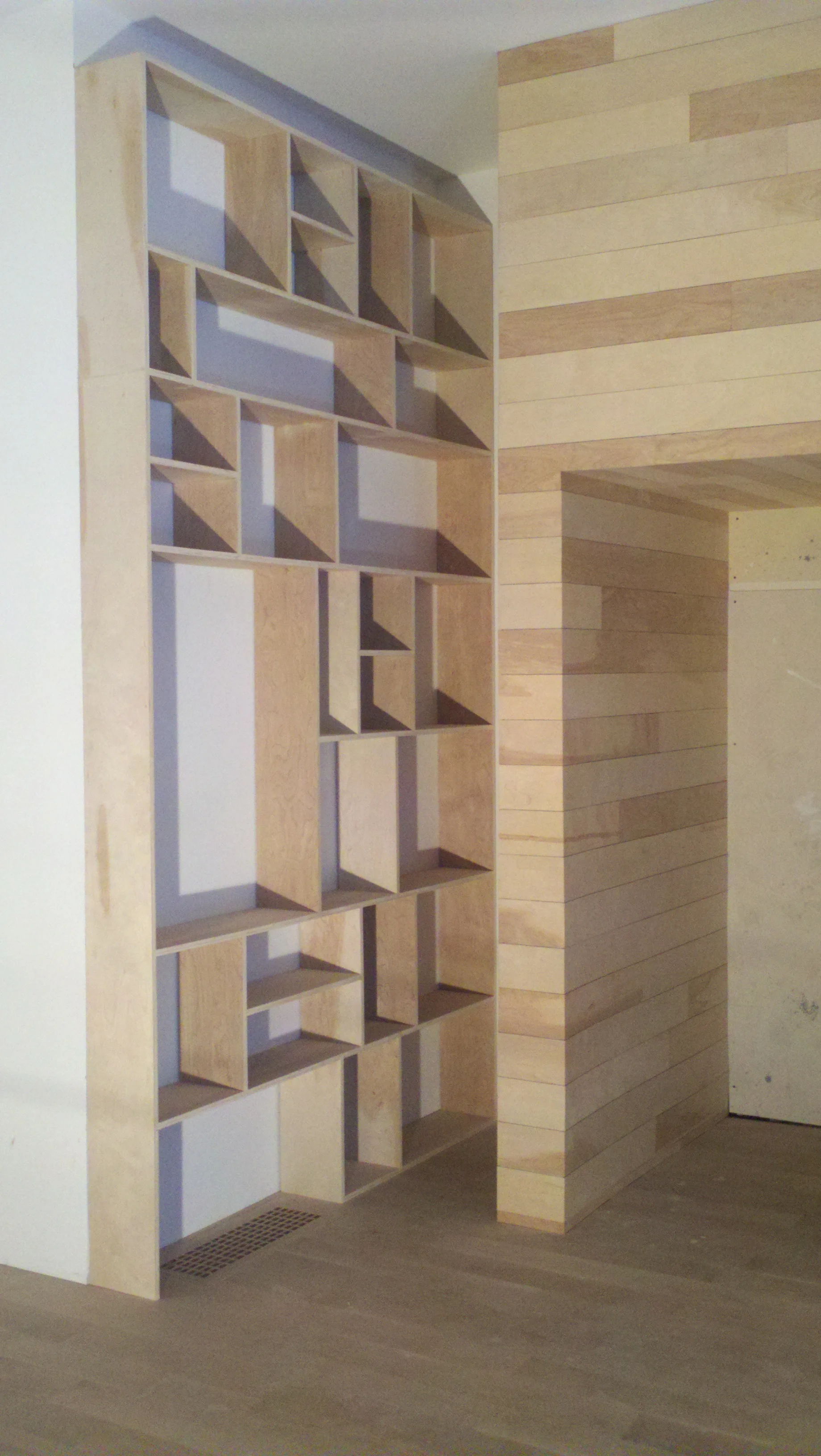 Wooden shelving unit with various sized compartments next to a curved wooden wall in a room with wooden floorboards.