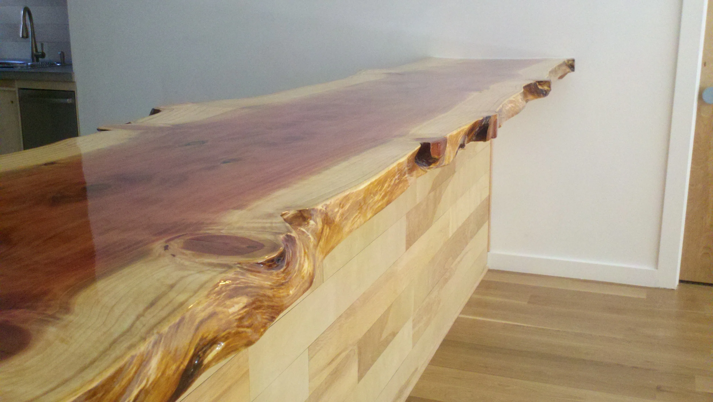 A polished, live edge wooden countertop or table in a room with wooden flooring and white walls, with a kitchen sink and faucet visible in the background.