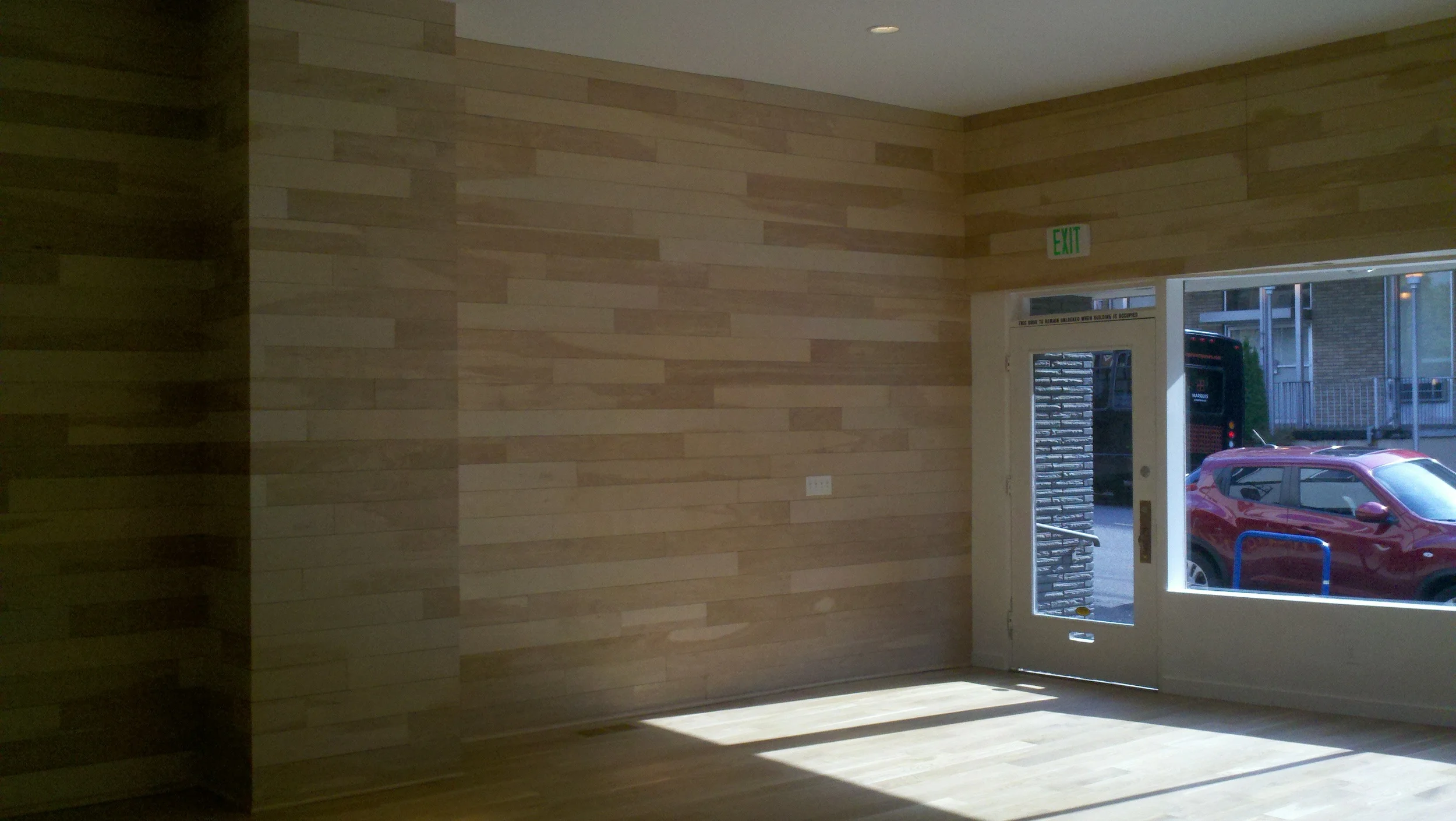 Interior of a room with wooden wall panels and a glass door leading outside, where a red car and a bus are visible.