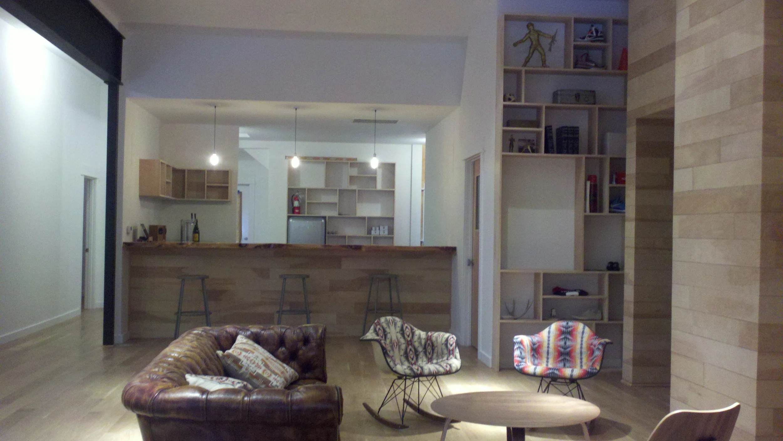 Living room with a brown leather sofa, patterned chairs, a round coffee table, and an open kitchen area with barstools, wooden shelves, and hanging pendant lights.