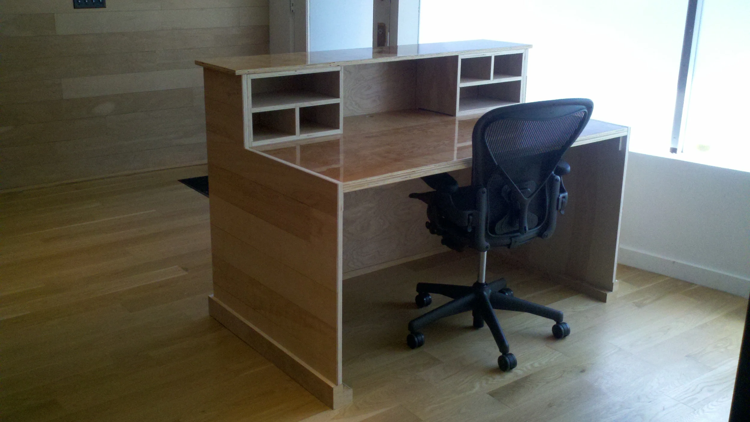 Wooden desk with cubby shelves and a black office chair in front of a window.