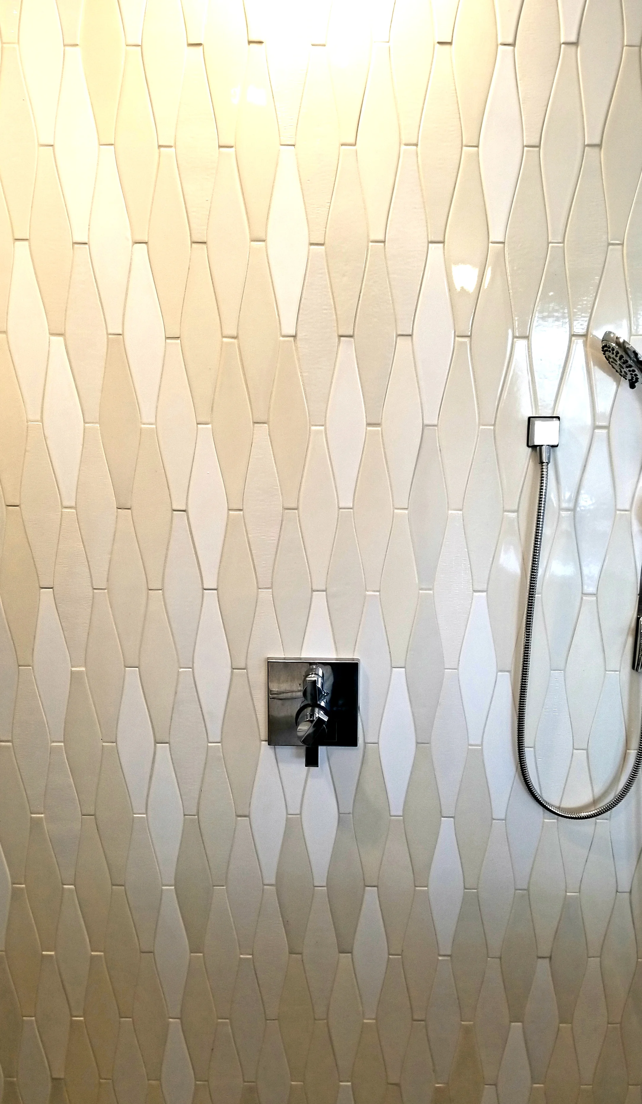 A tiled bathroom shower wall with a chrome shower handle, a metal square shower control, a flexible hose, and a showerhead.