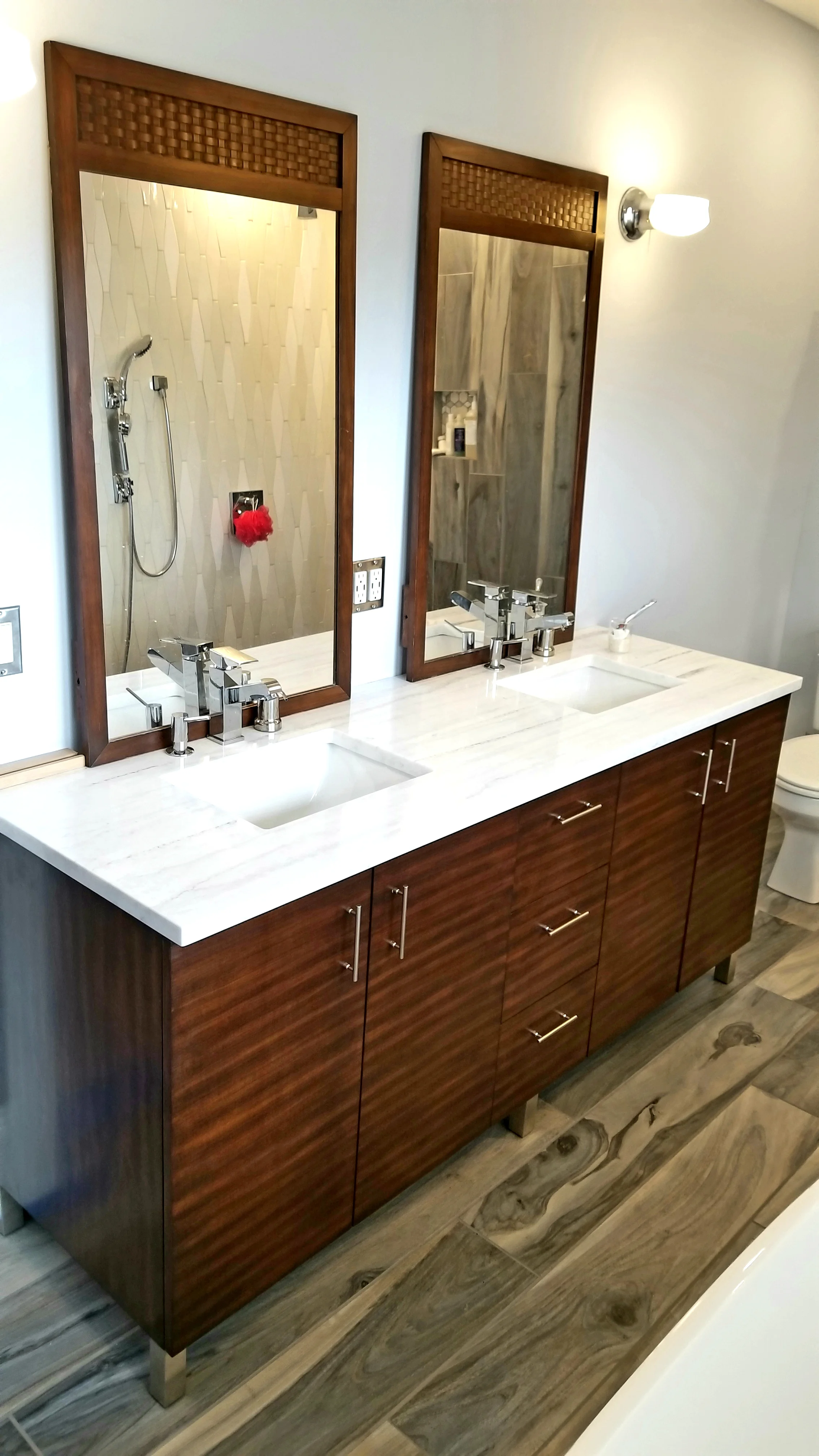 Double bathroom vanity with wood cabinets, white marble countertops, two sinks, two large mirrors with wooden frames, and modern chrome faucets. There is a wall-mounted light fixture above the right mirror, and a small cup with a toothbrush on the countertop. A shower area with beige textured tiles, a showerhead, and shampoo shelves is reflected in the left mirror.