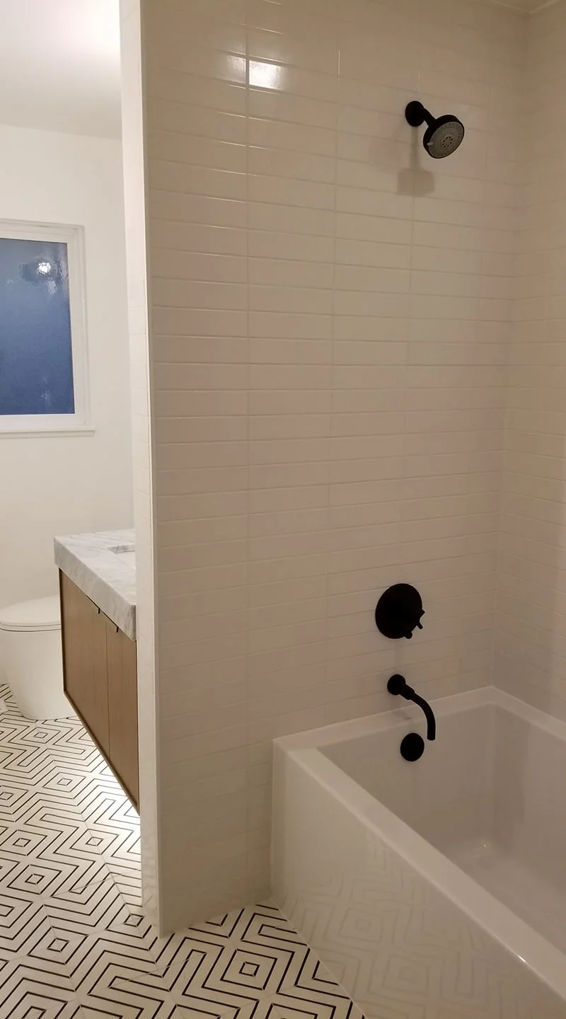 Bathroom with a bathtub, black fixtures including a showerhead, faucet, and handle, beige tiled walls, patterned black and white tile floor, and a partial view of a toilet and vanity with marble countertop.