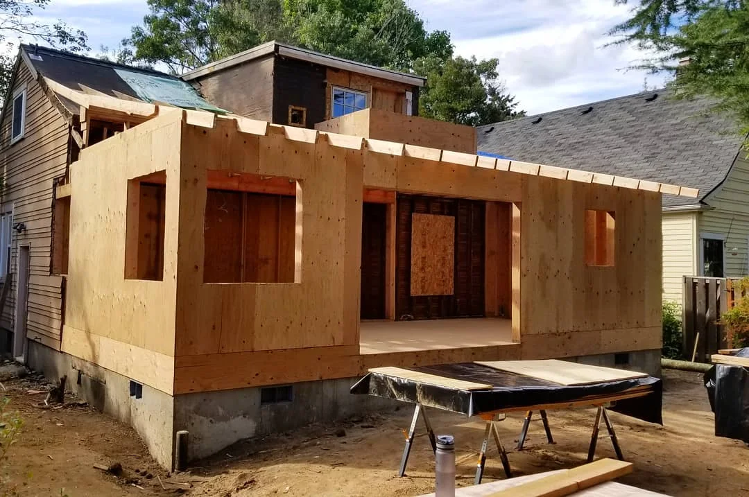 A house under construction, with a new wood frame and plywood walls. The build is in progress, with neighboring houses visible in the background.