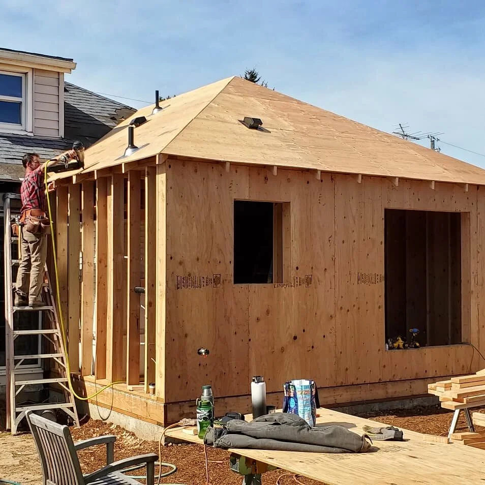 Wooden house under construction with a worker on a ladder installing roofing, outdoor furniture, and tools on a table.