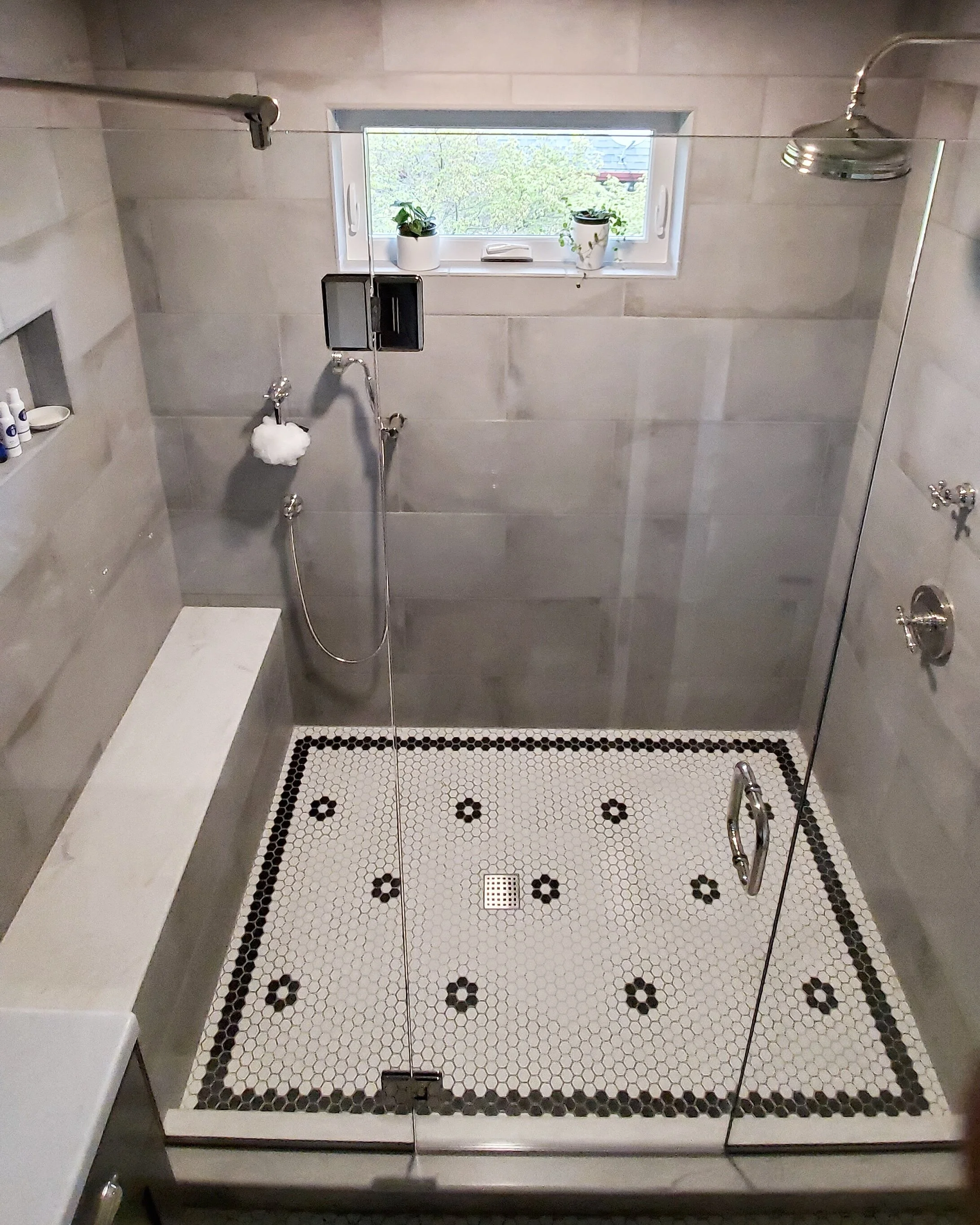 A bathroom shower with a glass door, featuring a black and white tiled floor with a geometric pattern, a window with potted plants, a handheld showerhead, and a shelf with toiletries.