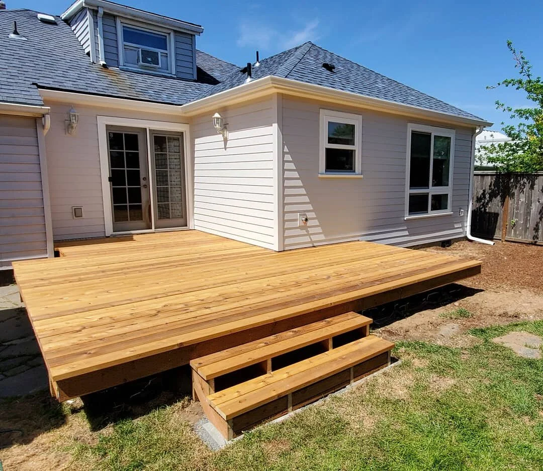 Newly built wooden deck attached to a white house with sliding glass doors, small windows, and a pitched roof.