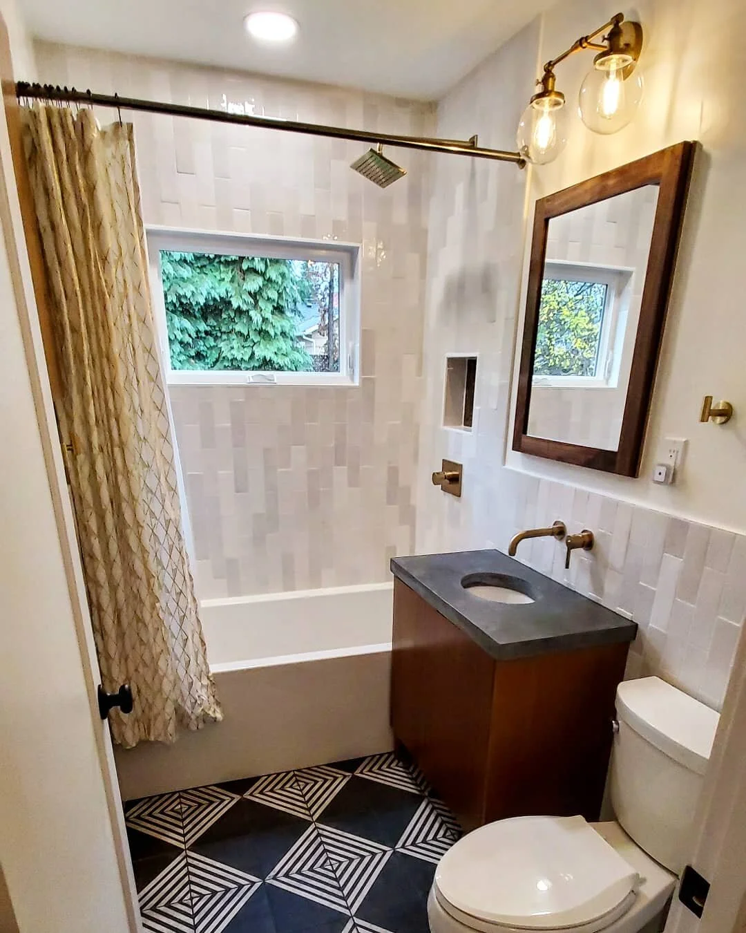A modern bathroom with a bathtub, window, shower head, vanity with sink, mirror, toilet, and black-and-white geometric floor tiles. The shower has a curtain and a built-in niche.