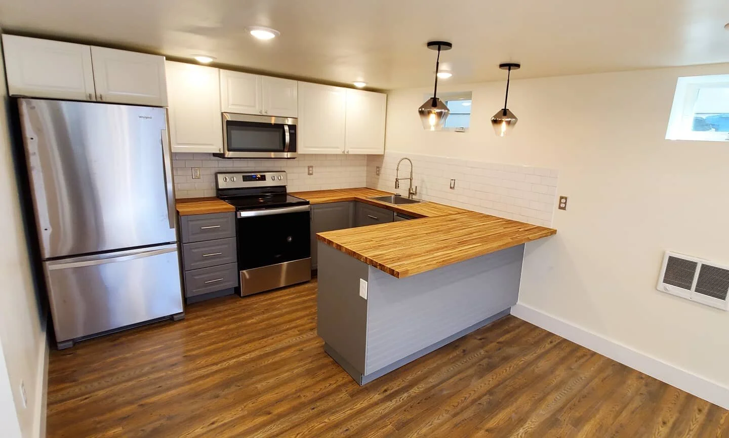 Modern kitchen with stainless steel refrigerator, black stove, microwave, gray cabinetry, wooden countertops, and wooden flooring.