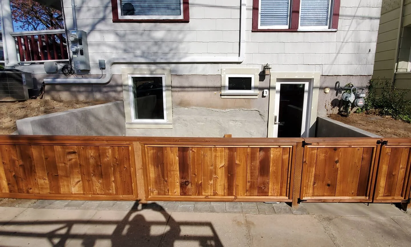 View of a backyard with a new wooden fence, concrete patio, and the back side of a house with two small windows and a door.