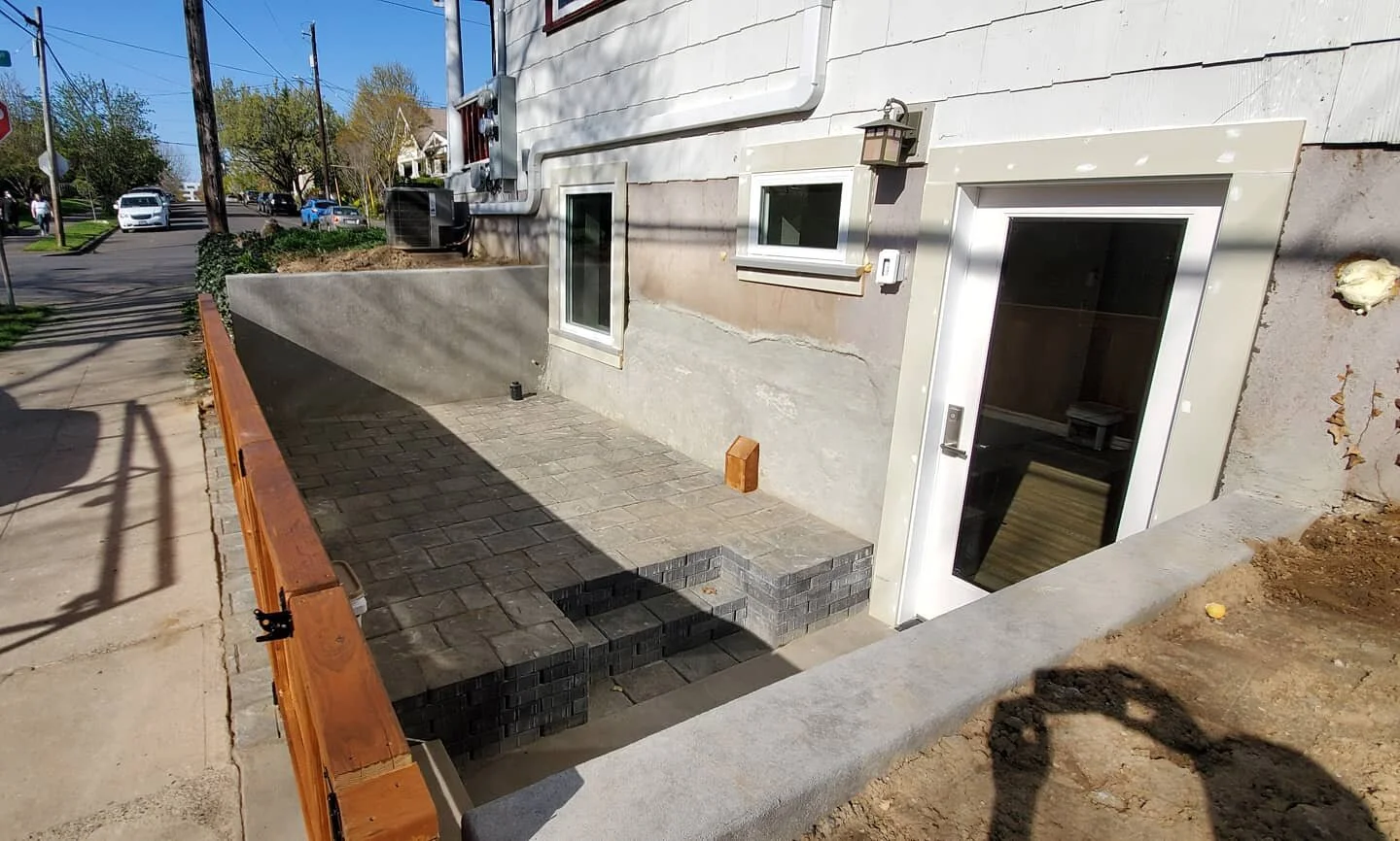 A back patio area under construction with stacked pavers, a small brick wall, a plastic chair, and a glass door leading into a building.